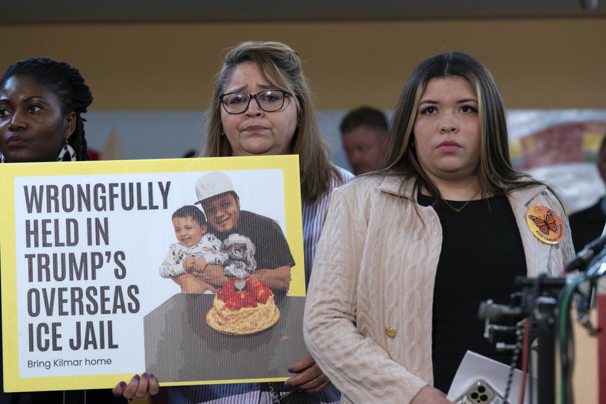 A woman stands at a microphone while another holds a poster reading "Wrongfully Held in Trump's Overseas ICE Jail."