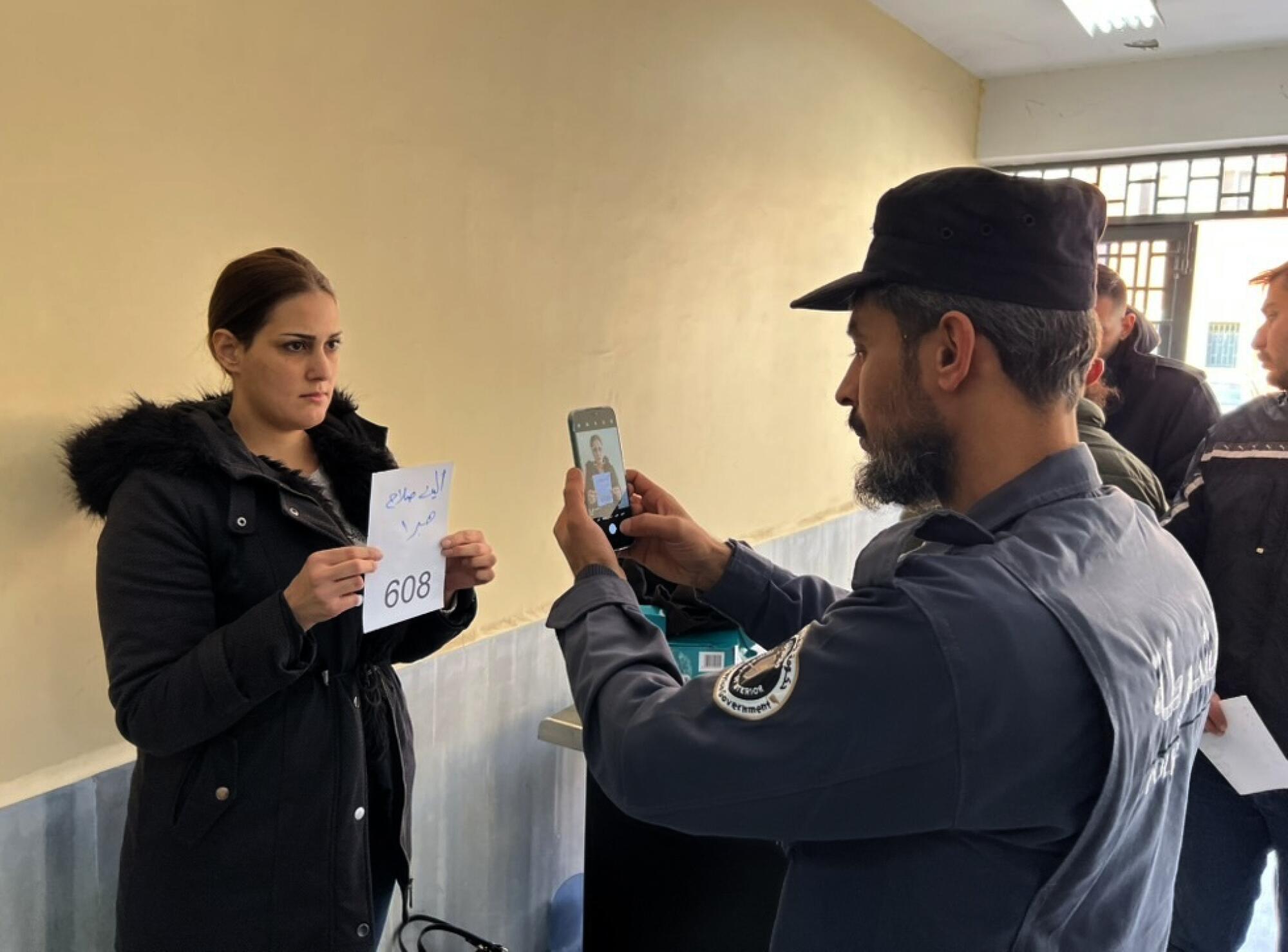 A man uses a cellphone to photograph a woman in dark clothes holding a white paper