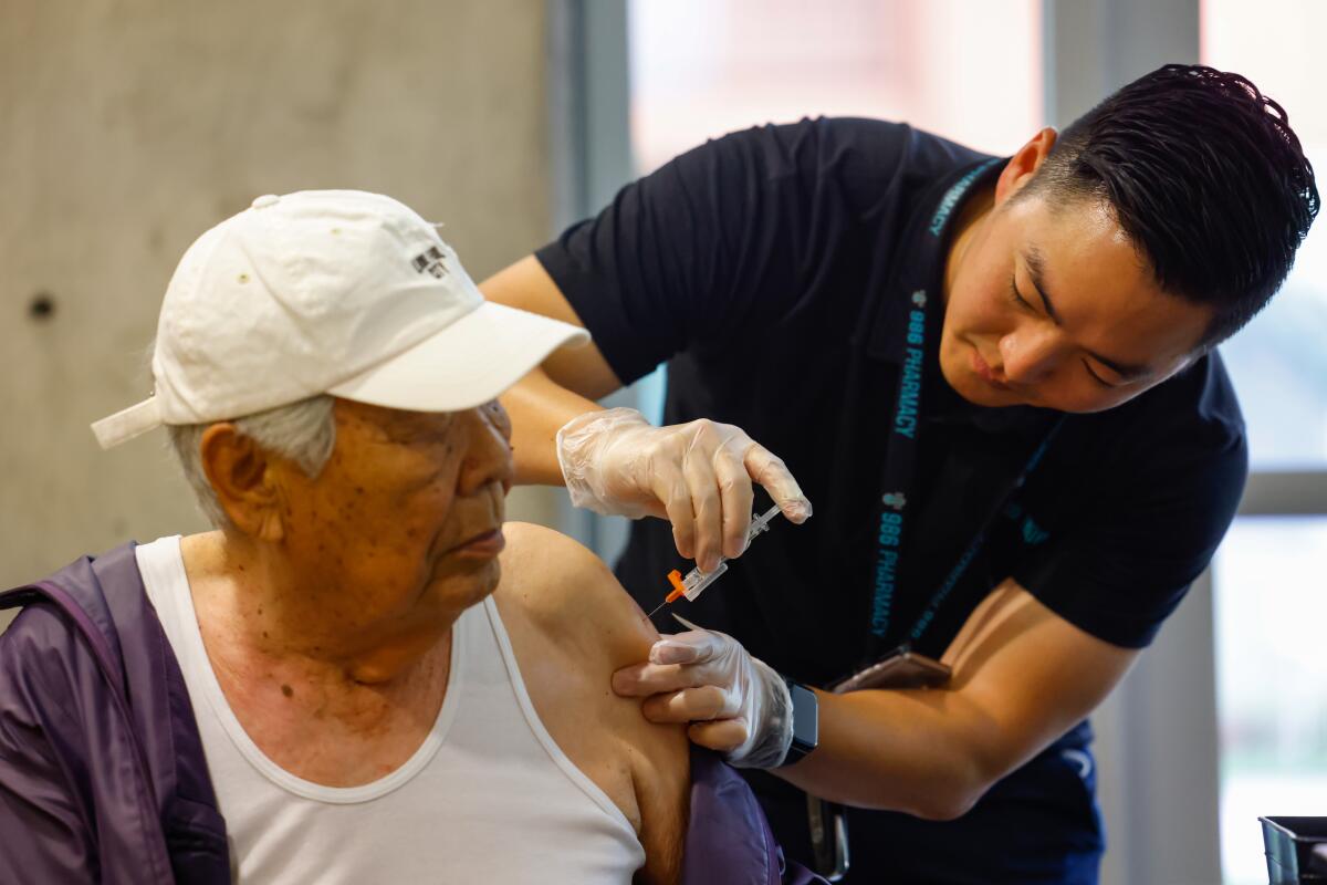 A man sitting with his left shirtsleeve pulled down as another man leans over and gives him an injection in the upper arm