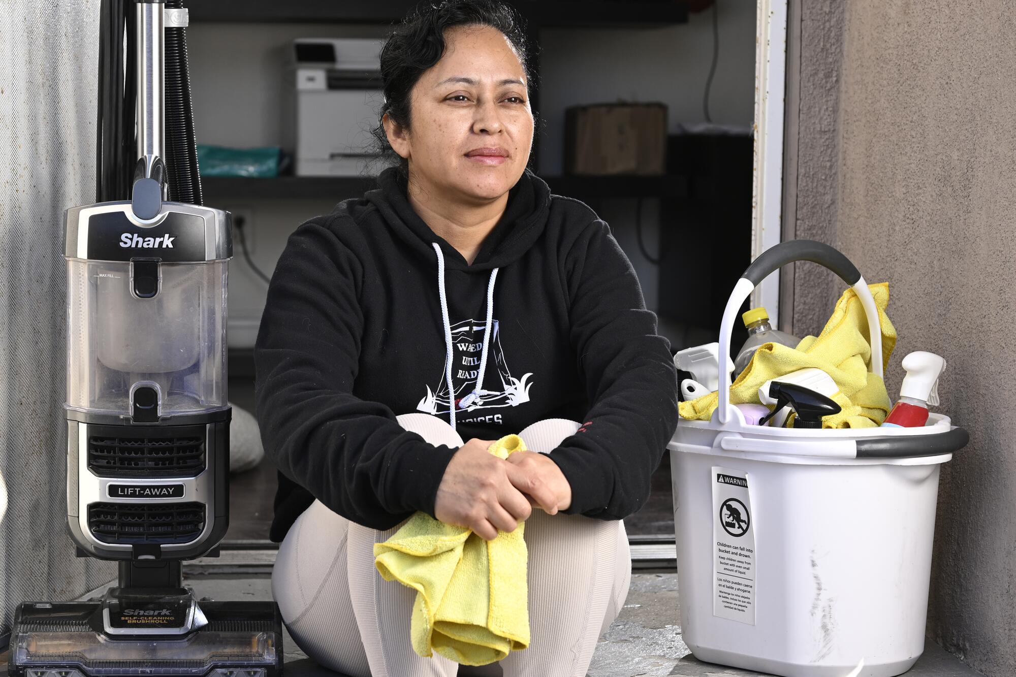 A woman with dark hair, in a dark sweatshirt and cream pants, sits holding yellow cloths next to a bucket with supplies