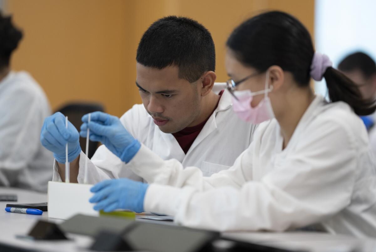Two people in white coats handle swabs at a desk.