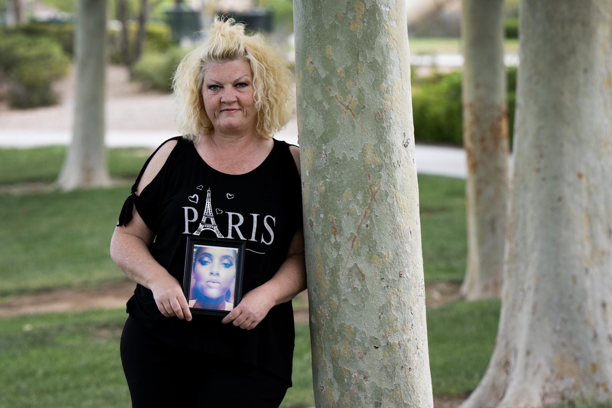 A woman leans against a tree trunk as she holds a photo of a younger woman.
