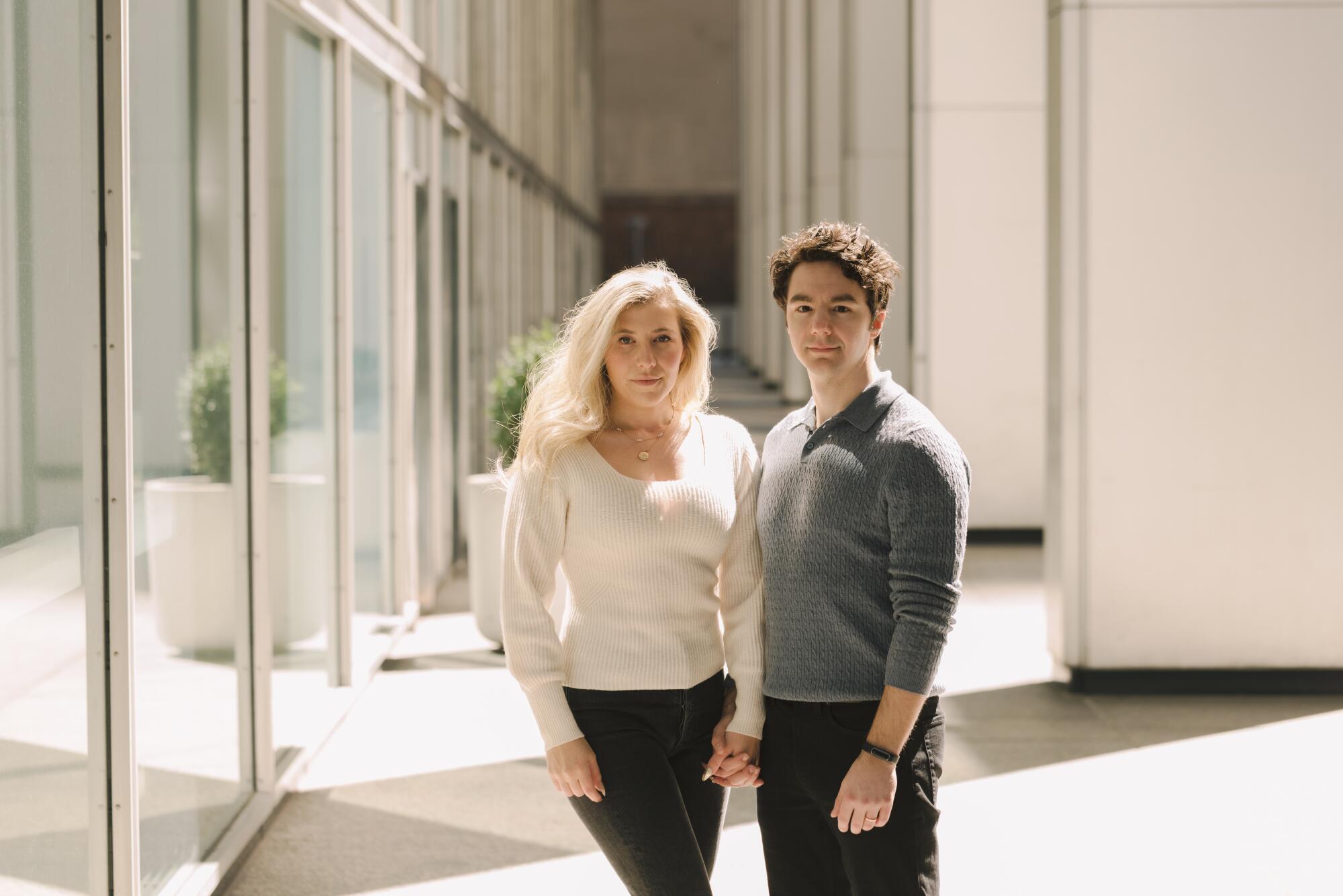 Linnea Sage and Paul Skye Lehrman stand together in a corridor lined with a wall of glass windows