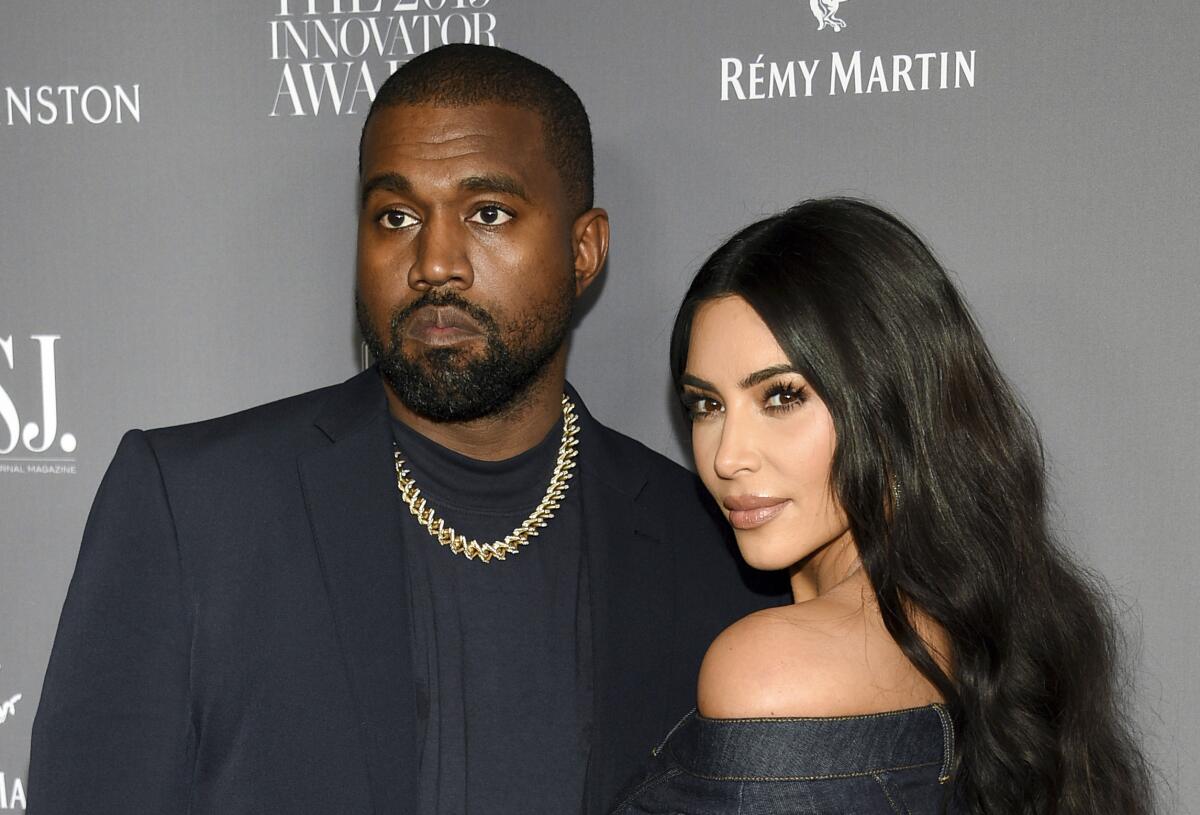 A man in a black blazer and gold chain poses with a woman in a black dress