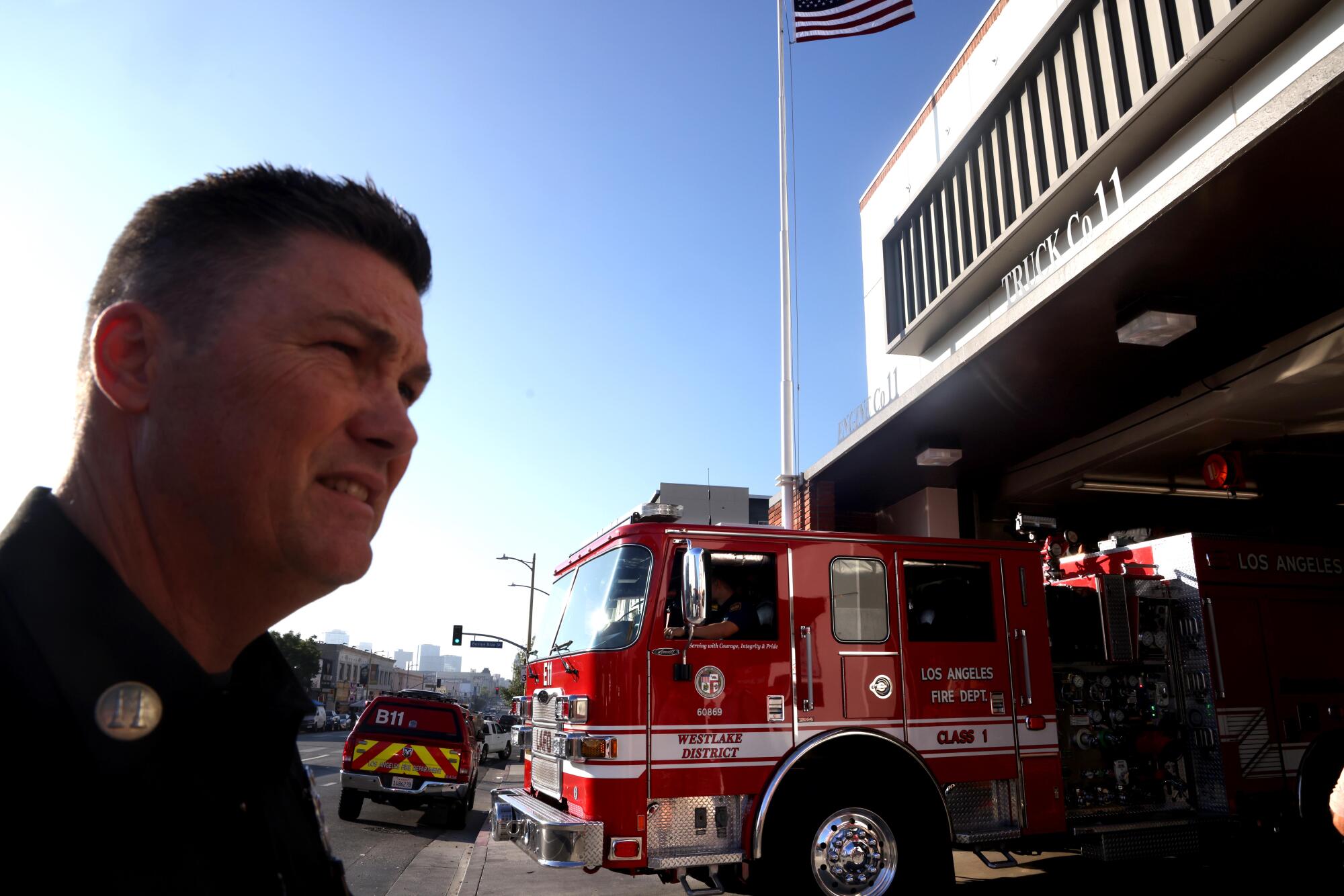 A fire captain stands outside a station as a truck departs.