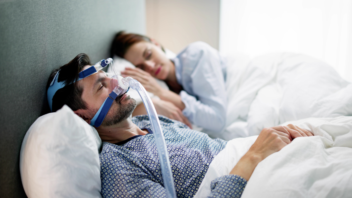 Obstructive Sleep Apnea Treatment of A photo of a man wearing sleep apnea oxygen mask equipment in bed with his partner.