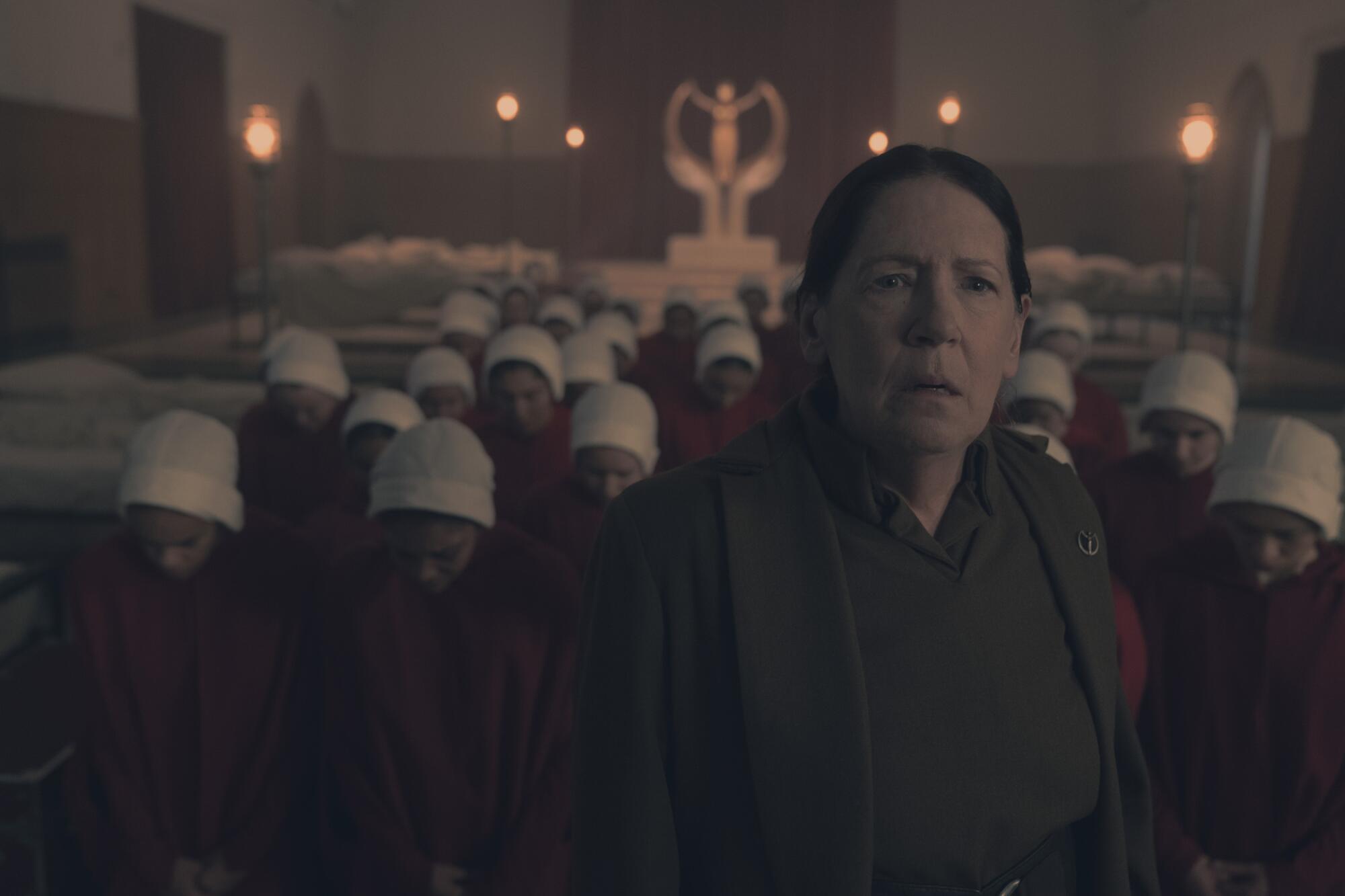 A woman in dark clothing stands in front of a group of women in red robes and white bonnets.