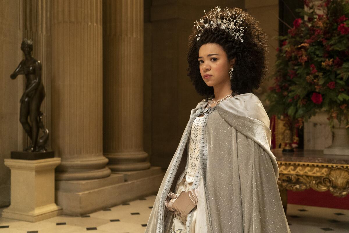 A woman with curly brown hair and a crown standing in a white dress and velvet cloak