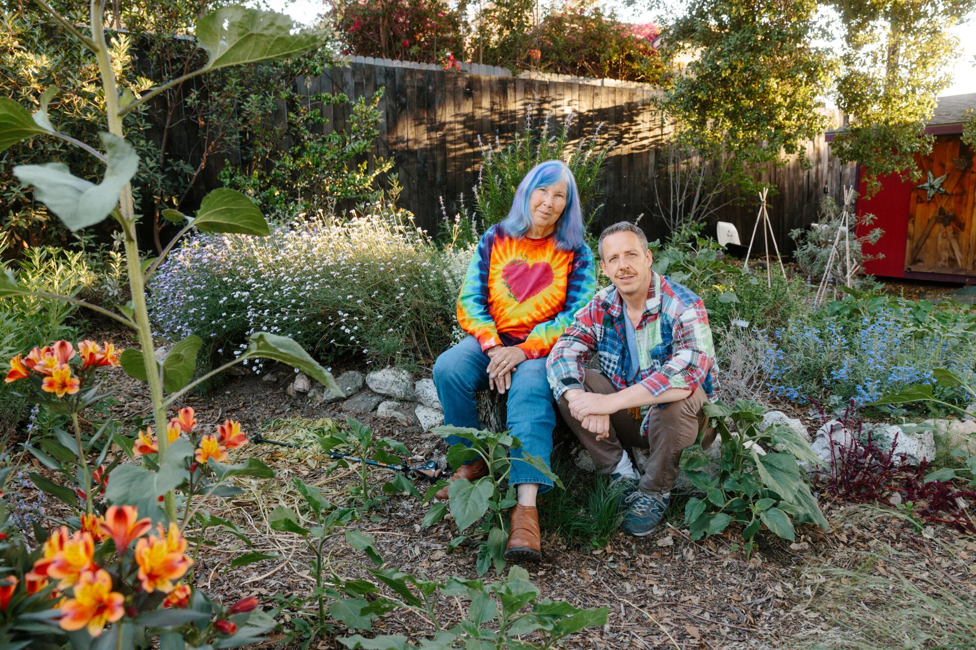 A woman with blue hair and tie-dye shirt sits in a flower garden next to a man wearing a quilted shirt of many colors.
