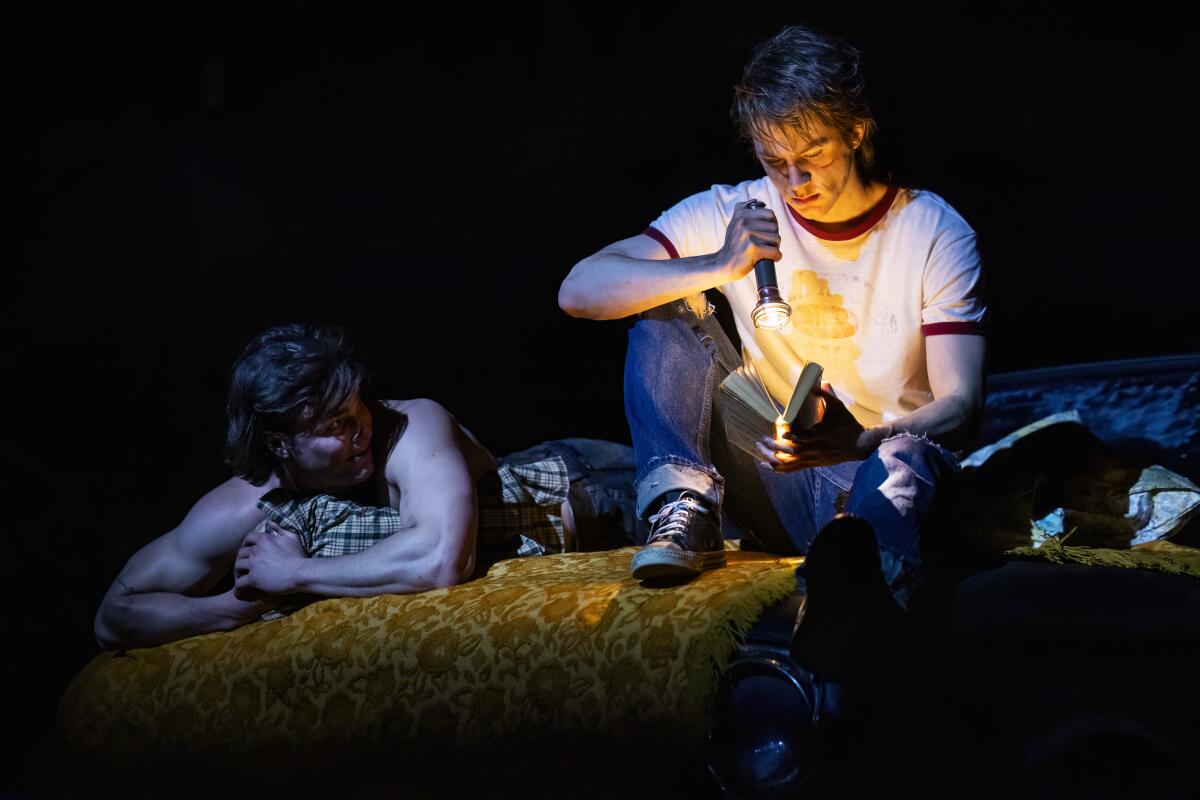 Two young men, one lying face down, one sitting on the ground looking at an open book with a flashlight